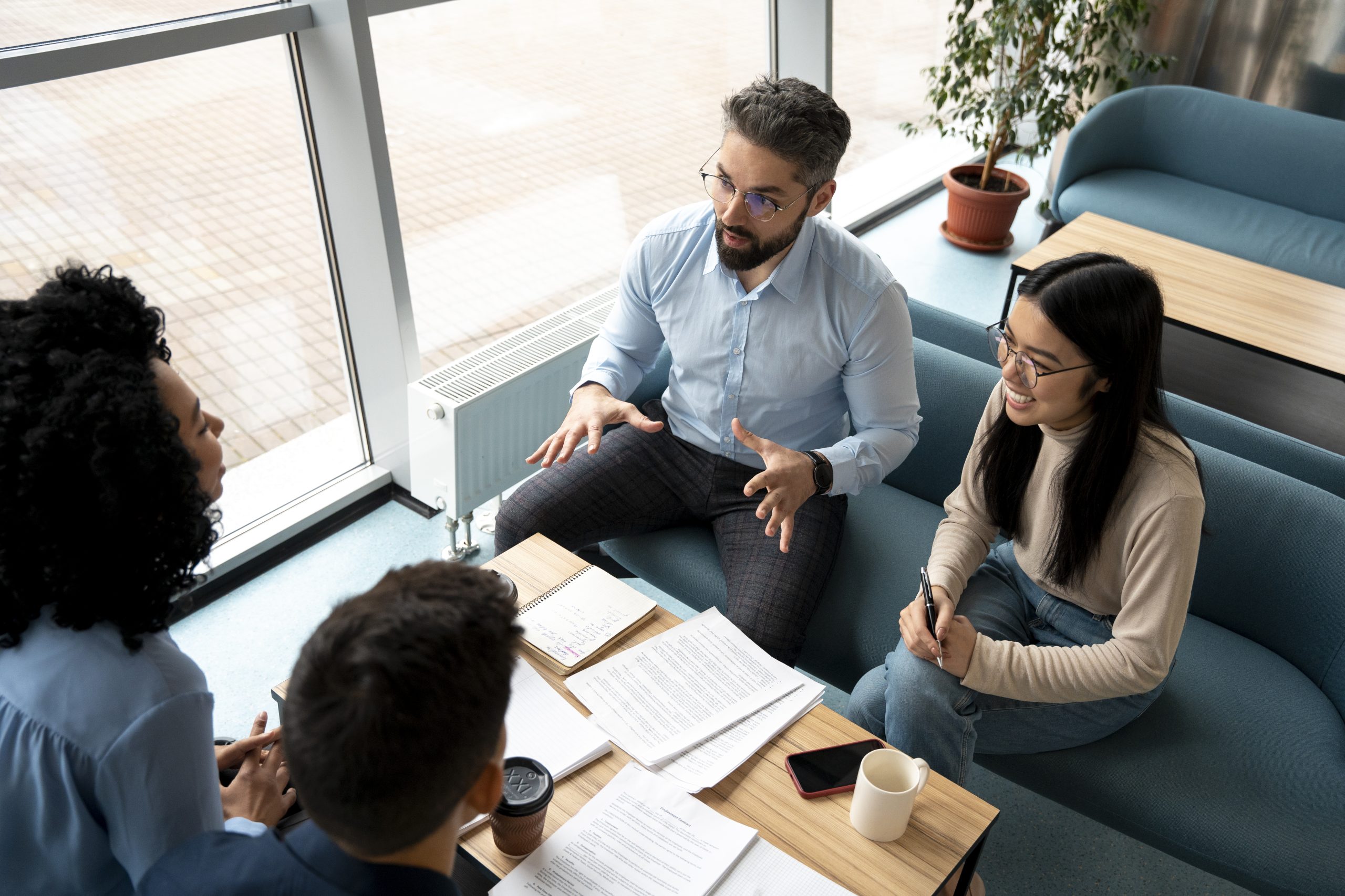 Quatre personnes en réunion informelle dans un espace de travail lumineux. Elles sont assises autour d’une table basse avec des documents, des carnets, des tasses et un téléphone portable. Un homme au centre parle en gesticulant, pendant que les autres l’écoutent attentivement.